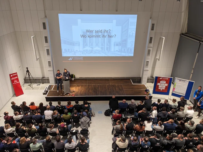 Eröffnung des Barcamps. Zwei Personen stehen auf der Bühne und präsentieren. Im Publikum ist gut gefüllt. 