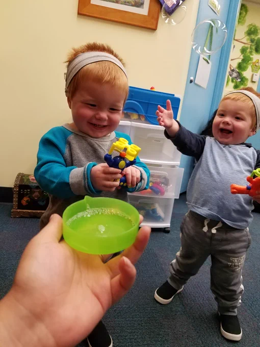 Two young toddlers in a play room. they each have short light hair and wearing a headband. They are holding a toy figurine. A person is holding a plastic round dish and extending it toward them. 