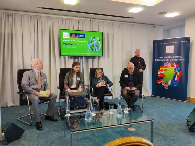 Picture of four seated people and one standing person. One seated person has a mobility device. Four seated people are panelists. One standing person is a sign language interpreter.