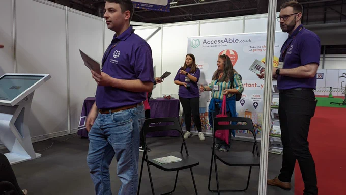 Two white men on the stand AccessAble are running at Naidex and one white woman talk to people visiting the stand about the company access guides