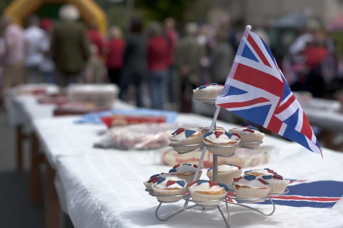 A stand of cakes with Union Jack flags sticking out of them on a long table and in the background people can be seen standing.