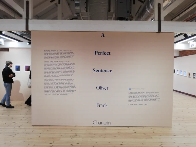 Down the middle of an exhibition wall, large text reads 'A Perfect Sentence' and 'Oliver Frank Chanarin' to each side there is more text about the exhibition but it's too small to read. You can see two people on the left who are looking at framed photographs on the wall.