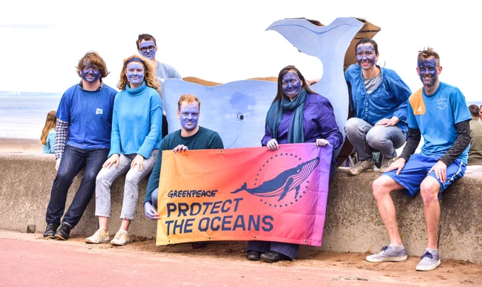 The Greenpeace Edinburgh team sits on a wall by the ocean, faces painted blue, holding an orange and red banner that says "Protect the Oceans". A large cardboard whale sits behind them.