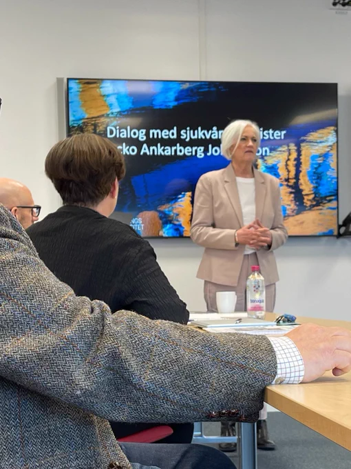 Sjukvårdsminister Acko Ankarberg Johansson står vid kortänden på ett bord i ett konferensrum. Bakom henne en digital skärm med texten "Dialog med sjukvårdsminister Acko Ankarberg Johansson"