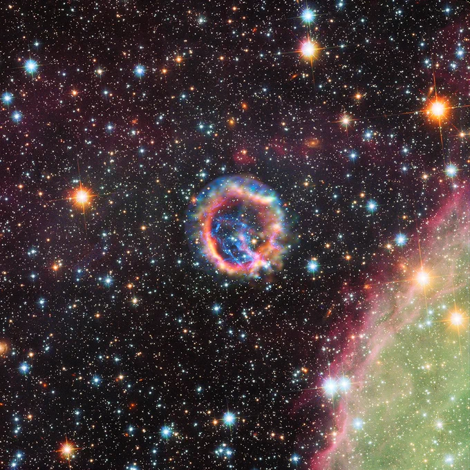 This is a composite image combining X-ray and optical light. Towards the center of the image is a fluffy ring that appears to contain every color in the rainbow. Starting with the innermost portion of the ring, the colors flow outward like mist in shades of red, then orange, then yellow, then green, blue, and purple. In the center of the ring there is a blue structure that resembles lightning. At the lower right of the image, a lime green cloud in the shape of an arc is present. Its inner edge is highlighted in shades of red and pink. Stars of all shapes, sizes, and distances are present throughout the entire image to dazzling effect.