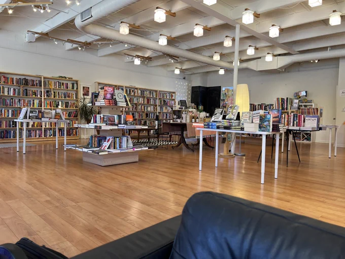 Wide shot of a bright and spacious indie bookstore, a rainbow of paperbacks in wall mounted shelves, and several display tables.