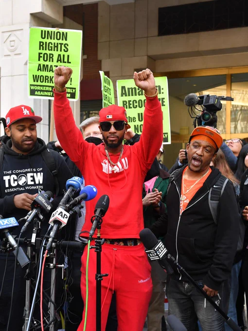 Chris Smalls wearing red Amazon Labor Union gear holds up two fists at a press conference next to his comrades from TCOEW The Congress of Essential Workers in front of many press mics. People gather behind him holding neon green signs that read: "UNION RIGHTS FOR ALL AMAZON WORKERS" and "AMAZON RECOGNIZE THE UNION NOW."