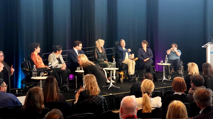 Seven people, five of which are in wheelchairs, are sitting on a stage in front of a dark backdrop. They are engaged in conversation.
