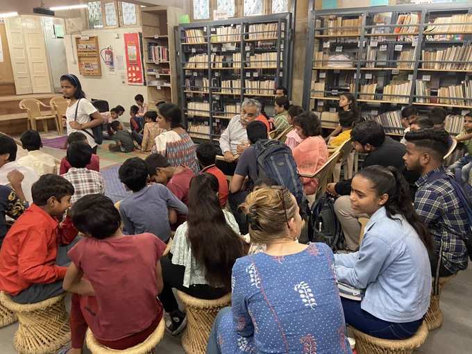 Image 3: inside the library, members and guests are seated on moodas and chatai before the program begins. They are surrounded by bookshelves