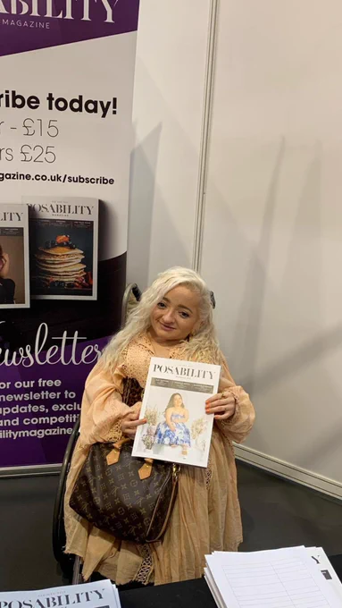 A picture of Samantha Renke inside PosAbility's booth at Naidex. She is using a wheelchair, and wearing a gorgeous wheat-coloured flowy dress, paired with a dark brown Louis Vuitton bag under her arm; her white blonde hair spills over her shoulders. She's smiling and holding a copy of PosAbility Magazine, showing a short stature woman in a white and blue dress.