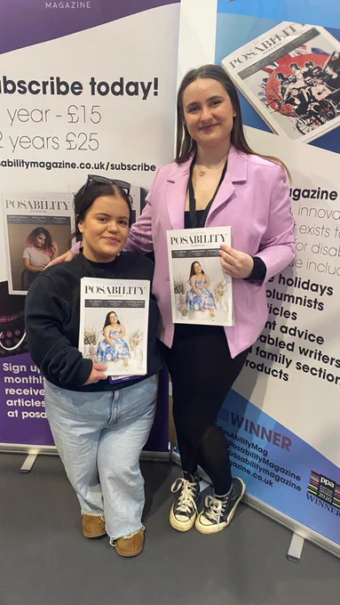 Two people in front of large advertisements for PosAbility Magazine. On the left is a woman of short stature with her hair tied back, wearing a dark baggy jumper and light jeans. On the right is a woman in a bright pink blazer, black leggings and converse. They are both holding copies of PosAbility Magazine that show the woman on the left in a white and blue dress, and are smiling at the camera.