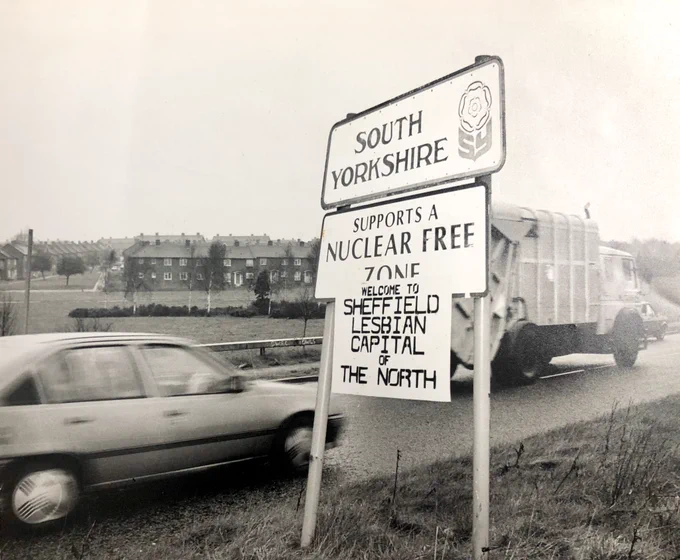 Old black and white photo of a road with cars going past, there is a road sign that says ‘south Yorkshire’ below it is another sign that says ‘supports a nuclear free zone’ and below that someone has added their own makeshift sign that reads ‘welcome to Sheffield lesbian capital of the north’