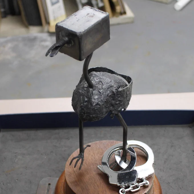 A picture of a metal bird statuette called 'The Thrang' on wooden base with metal handcuffs fixed to left leg.