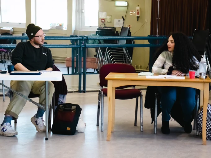 Photo by Lucy Smith Jones. Two people sit at desks and look over, talking to each other. On the desks are scripts and papers.