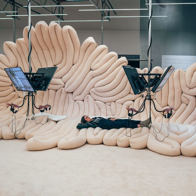 A photo of a female visitor lying on her back on the sausage pillow. She is wearing headphones and watching the screen in front of her.