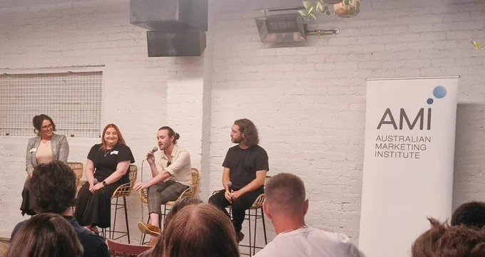 Four people sitting in high stools in front of a white brick wall during a panel discussion, next to a sign that says AMI. One person is speaking into a microphone. 