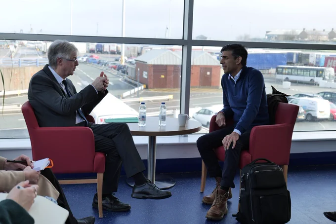 First Minister Mark Drakeford and Prime Minister Rishi Sunak having a meeting in Holyhead Port. The first Minister is explaining something in detail and the ports can be seen through the window behind them.