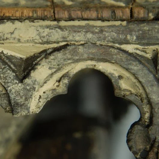 A wooden arc with carved details is covered with thick deposits of dirt. The surface of the object itself is dark while the dirt has a very light colour and covers large parts of the surface.