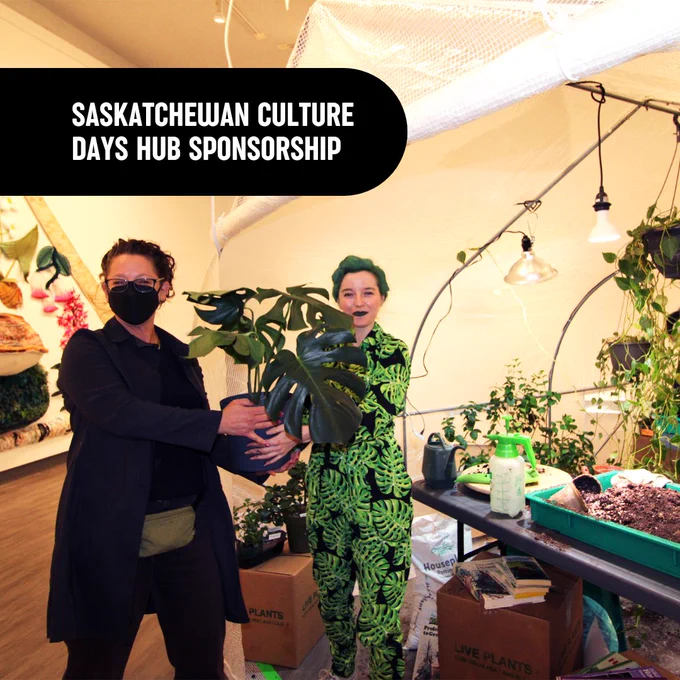 Plant Adoption by Artist Alyssa Ellis.
The Artist stands under a tent in the gallery space, filled with plants, wearing a plant pattern jumpsuit and green lipstick. The artist is handing a large monstera plant to a participant. 