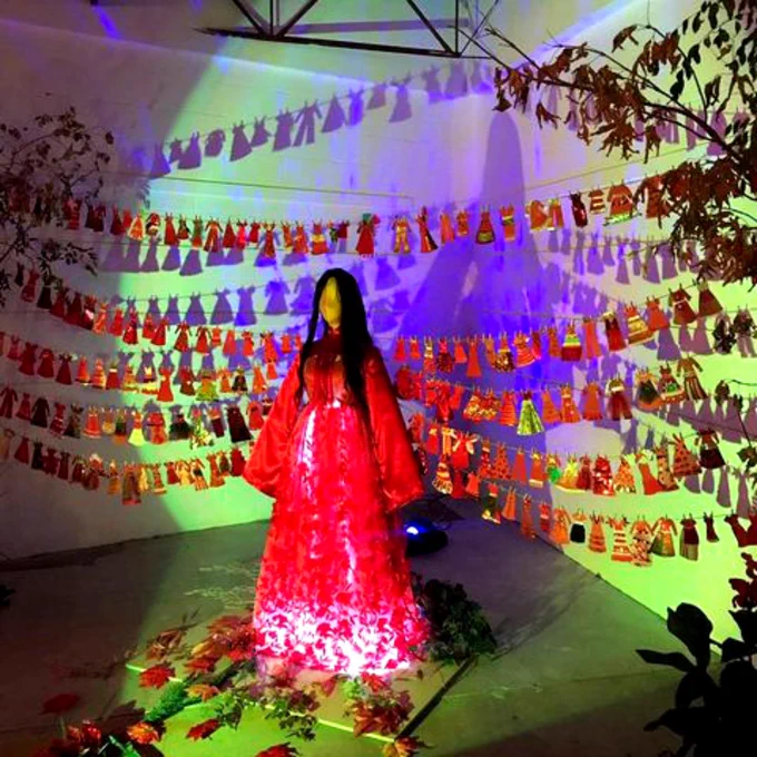 April Doepker's art piece The Many Red Dresses for Awareness Project features a mannequin wearing a red dress lit from below with a spotlight. There are strings hanging behind it displaying many small paper red dresses made by participants. 