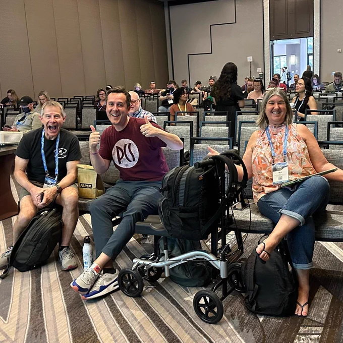 Two men and a woman, big grins at the camera, seated in the front row of a convention center room.