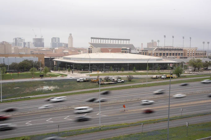 A view of the UT football stadium in the background with buildings to either side, including the UT tower to the left. In the foreground is a highway with blurry cars driving past. Photo by Alex L.