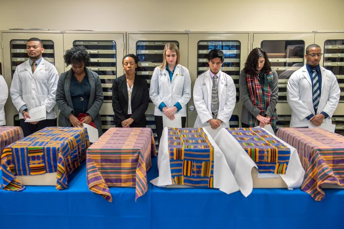 Photograph of people (some in formal clothes, some in lab coats) bowing their heads over boxes covered in brightly patterned cloth. 
