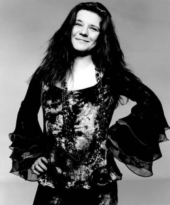 Fotografía en blanco y negro de una sonriente Janis Joplin en 1970. Con el pelo suelto, un largo collar y una blusa de anchas mangas, exhibe una media sonrisa entrañable. Foto de Grossman Glotzer Management Corporation. Dominio público.