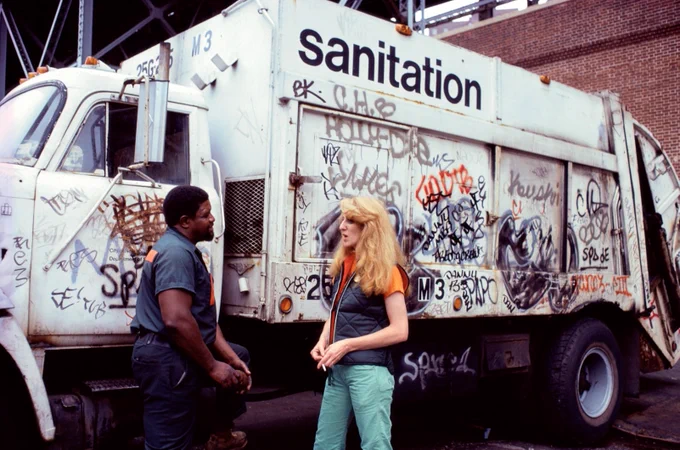Ukeles speaks to a sanitation worker in front of a sanitation truck. The truck is white and covered in graffiti 