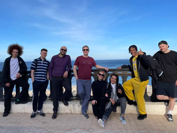 Cat sheridan, Billy Reid, Warren Murray, Ben Randall, Kameel Myrie, Neil Webb, Humza Koudri and Wahiba Ghanem leaning against a railing with blue sea and sky behind them