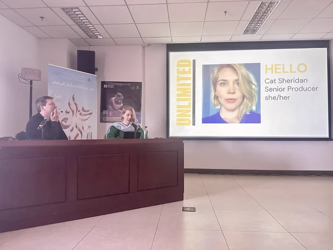 Cat Sheridan, senior producer for Unlimited and a Neil Webb, Director of Theatre and Dance for the British council - sitting behind a dark brown desk whilst speaking on to microphones and presenting next to a PowerPoint projection