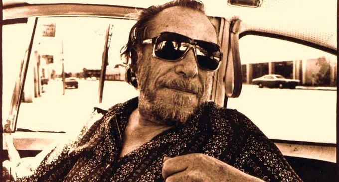 Charles Bukowski wearing sunglasses behind the wheel of his car.