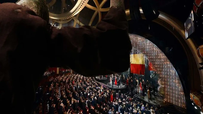 A hunched over figure in the rafters above a full Dolby Theatre.