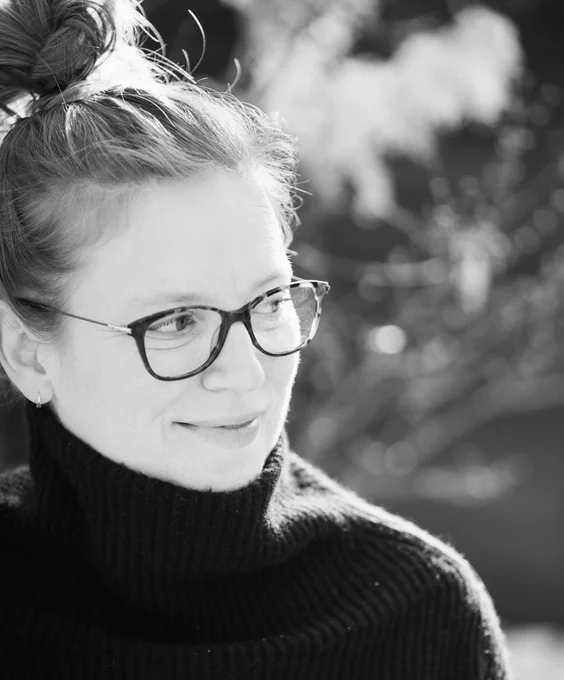 A black and white portrait of filmmaker Sarah Polley. She wears her hair in a bun, a black turtleneck, and dark-framed glasses, smiling slightly to the side. 