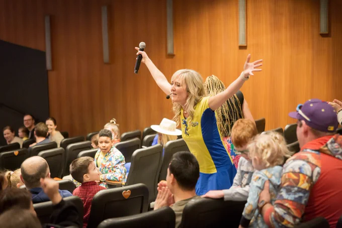 A woman with her arms raised stands in the middle of a theatre filled with children and their parents.