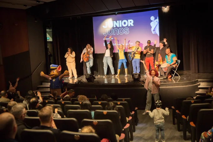 One child dances in the aisle as other young music fans and their parents sit in a theatre and watch musicians play guitar or raise their arms on stage.