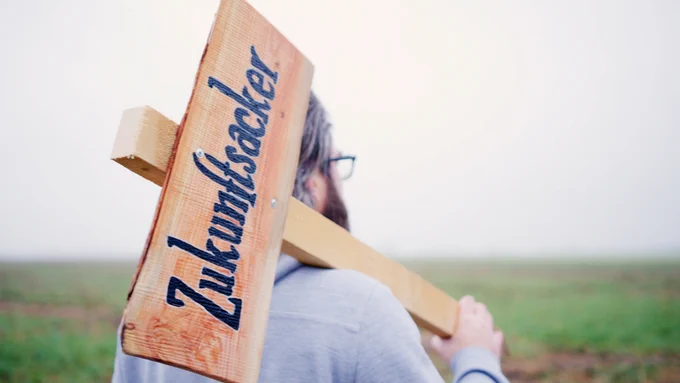 Auf dem Bild läuft @teraspri über ein Feld und trägt ein Schild auf dem Zukunftsacker steht. Es ist das Vorschaubild für das Crowdfunding auf Startnext: https://www.startnext.com/zukunftsacker