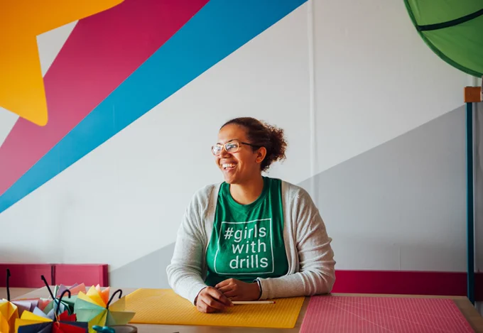 A woman sitting at a table, with yellow and pink cutting mats in from of her. She wears a beige cardigan and a green t shirt that reads #GirlsWithDrills. She is smiling and looking off to the left of the camera. The wall behind her is painted with a bright yellow star, pink, blue, white and grey stripes. 