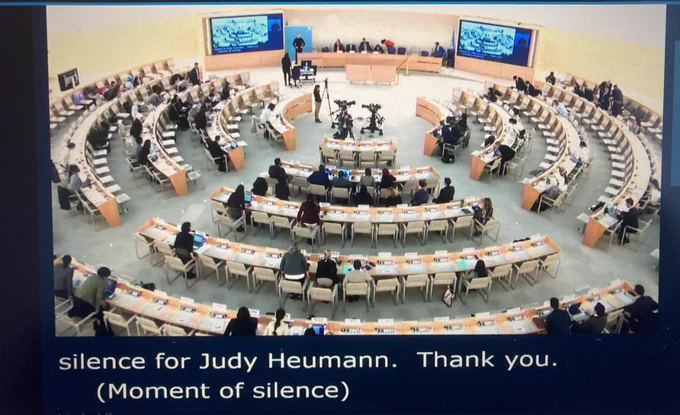 Wide shot of UN Human Rights Council chamber and those present standing in silence remembering Judy Heumann.