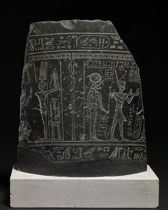 A large black stone fragment engraved with three Egyptian figures. The figure on the left wears a crown and makes offering to the other two, who are gods.