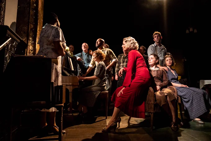 The residents of the boarding house gather around a piano, listening.
