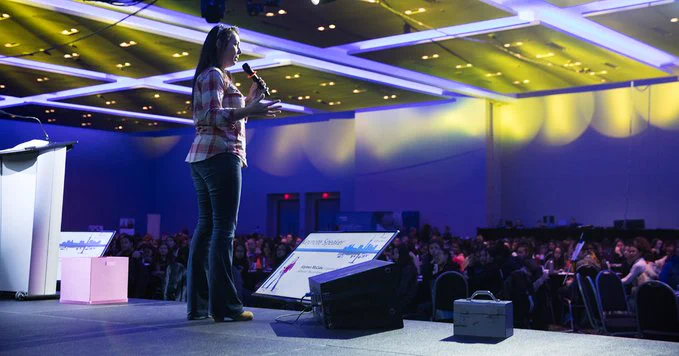 A skilled trades professional stands on stage while addressing a seated crowd in a conference room at a previous Young Women's Conference.