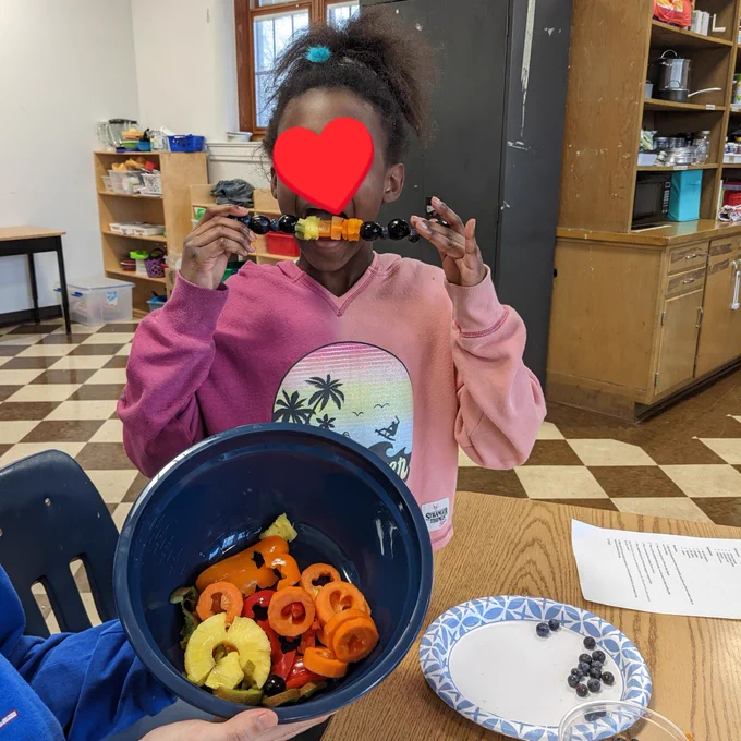 Young girl eating fruit skewer with bowl of fruit held in front of her. Standing in a classroom at a desk.