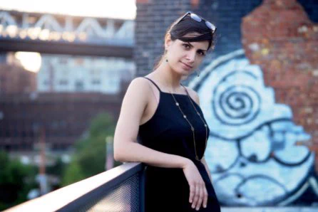 A professional headshot of Niloufar Nourbakhsh.  She is outside leaning against a metal railing.  She wears a dark dress with a high, square neckline and thin straps.  She also wears a dangling necklace and earrings.  Her hair is dark and tied back, and her head is tilted slightly toward the camera.  A pair of glasses sits on top of her head.  In the background, a brick wall is visible with graffiti-like designs covering portions of it.  Other buildings and tree tops are also visible on the other side of the railing.