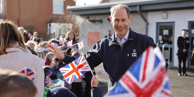 The Duke, smiling and waving to a crowd of people waving flags. 