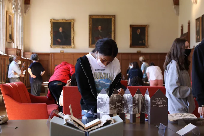 A young black woman looking at an artists' book titled "What's Happening with Momma." She is standing at a table where a number of other books are displayed. 
