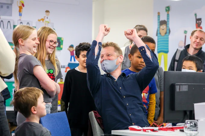 Young people and a teacher sitting at a computer celebrating at Raspberry Pi Foundation HQ.