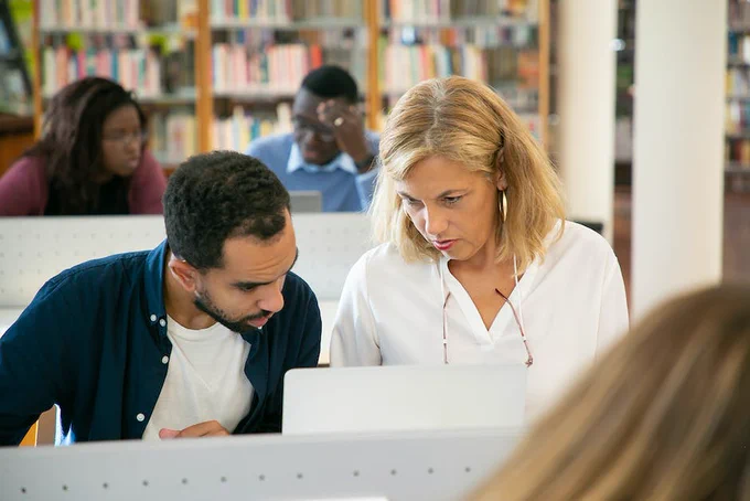 image depicts 4 individuals using a digital device in a library setting, in the foreground is a partially obstructed head of hair 
