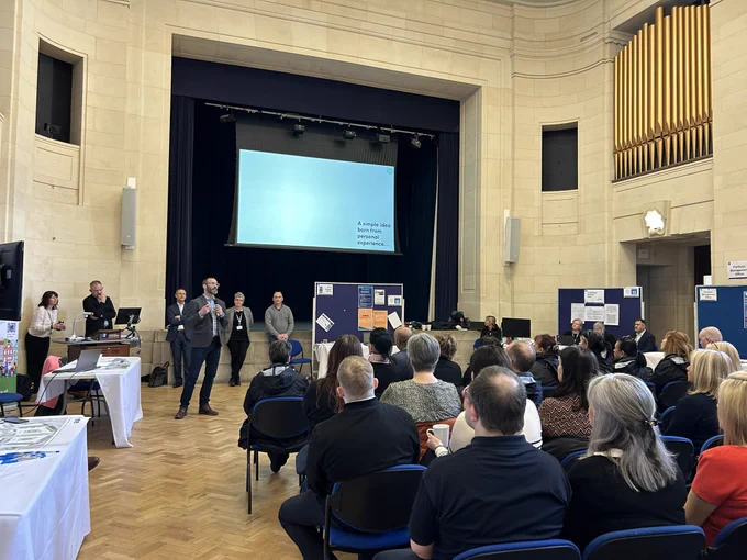 The great hall, Trent Building, packed audience watching AccessAble’s Director David Livermore deliver a presentation on the AccessAble university of Nottingham project. Information stalls can be seen around the perimeter of the hall.