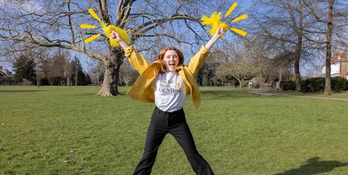 Girl with red hair does a starjump in front of camera. She is holding yellow pompoms, and wearing a yellow coat. She is in a park on a bright day. 